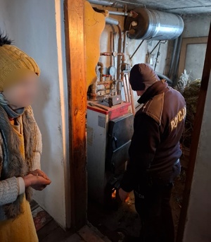 W scenerii domowej kotłowni policjant oraz seniorka przy piecu CO