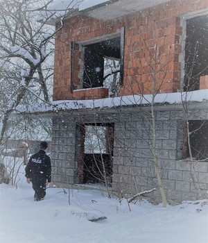 Policjant obok opuszczonego budynku mieszkalnego bez okien stojący w głębokim śniegu