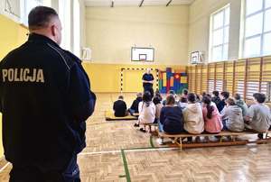 Dwaj policjanci w trakcie pogadanki dla dzieci siedzących na ławkach w sali gimanstycznej