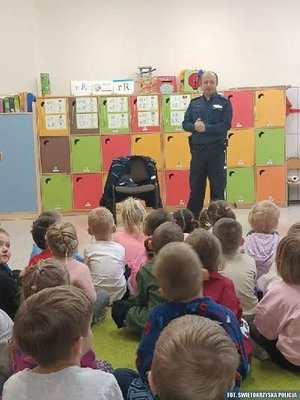 Policjant podczas pogadanki dla dzieci siedzących na wykładzinie sali przedszkolnej