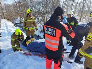 Grupa strażaków, ratowników medycznych i policjantów w scenerii zimowego potoku, na noszach osoba owinięta folią atermiczną