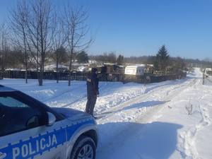W scenerii zimowej policjant obok radiowozu na tle ogródków działkowych
