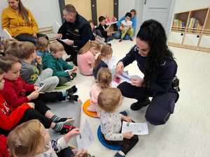 Policjantka oraz strażnik miejski pochylający się nad grupą dzieci siedzących na podłodze sali przedszkolnej
