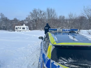 Odkryty ośnieżony teren, w oddali przyczepa campingowa, na pierwszym planie radiowóz i policjant