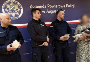 Na tle ścianki Komendy Powiatowej Policji w Augustowie trzej policjanci i kobieta z niemowlakiem na rękach
