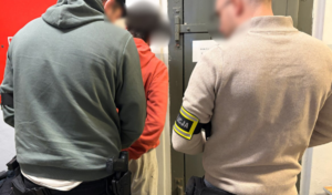 Widziani z tyłu dwaj policjanci po cywilnemu wprowadzający mężczyznę w czerwonej bluzie do pomieszczenia dla osób zatrzymanych