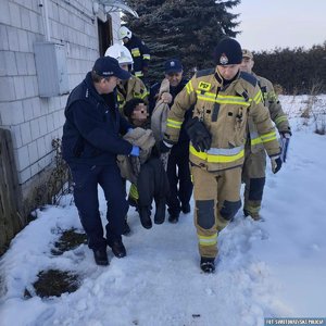 Policjanci i strażacy niosący na kocu staruszkę wzdłuż budynku