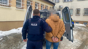 Widziani z tyłu policjant wprowadzający do radiowozu typu bus mężczyznę w kajdankach
