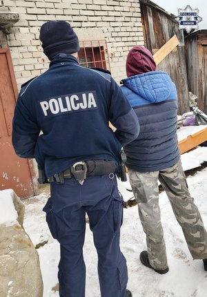 Widziani z tyłu na tle ruin budynków gospodarczych policjant i osoba w bluzie z kapturem
