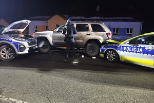 W scenerii zimowej nocy na jezdni samochód terenowy zablokowany przez dwa radiowozy. Jeden radiowóz ma uszkodzoną przednią część nadwozia.