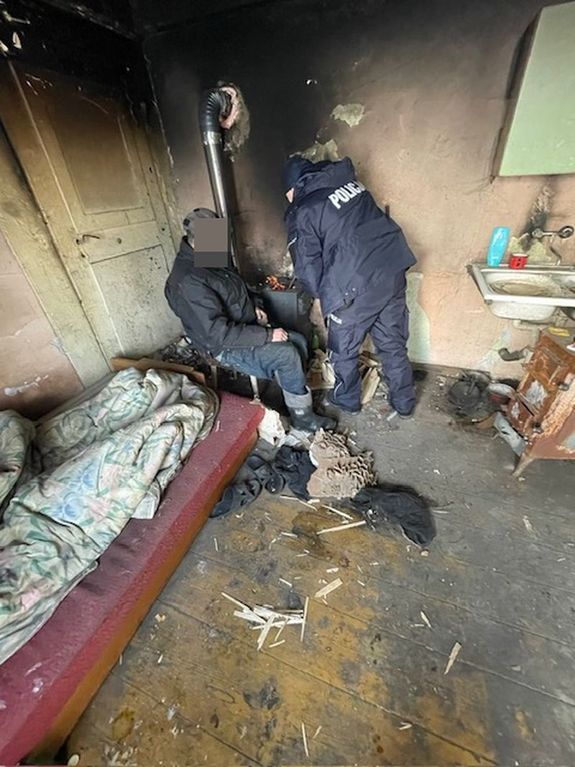 W zaniedbanym pomieszczeniu starszy mężczyzna obok pieca typu koza obok policjant