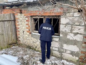 W scenerii zimowej policjant zaglądający przez wybite okno do wnętrz budynku gospodarczego