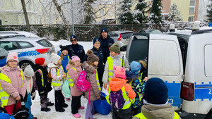 W scenerii zimowego dziedzińca komisariatu trójka policjantów, oraz grupa dzieci oglądających radiowóz