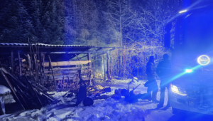 W scenerii  nocno-zimowej wóz strażacki i elementy drewniane przy budynkach gospodarczych