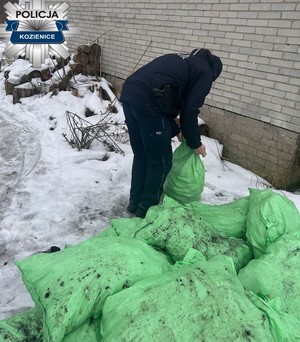Przy ścianie budynku policjant trzymający worek jutowy przy pryzmie worków