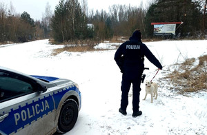 W scenerii ośnieżonego lasu radiowóz oraz policjant trzymający na smyczy białego psa