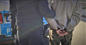 Zbliżenie pleców mężczyzny ze skutymi kajdankami rękami oraz policjanta na tle radiowozu.