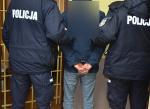 Widziani od tyłu dwaj policjanci i mężczyzna po cywilnemu przed kratami aresztu