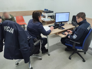 Dwójka policjantów przy stanowisku komputerowym, w tle mężczyzna ze zanimizowaną twarzą
