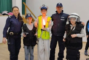 Dwójka policjantów oraz trójka uśmiechniętych dzieci z elementami policyjnego umundurowania pozujący do zdjęcia wewnątrz sali