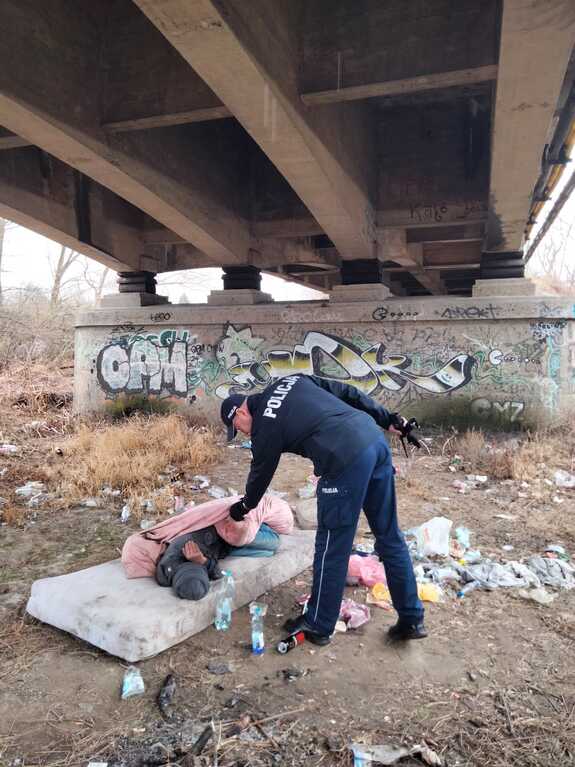 Policjant pochylający się nad bezdomnym mężczyzną leżącym pod mostem