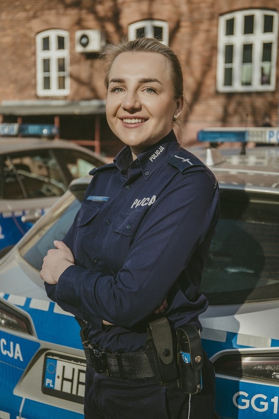 Uśmiechnięta policjantka na tle radiowozu i budynku z czerwonej cegły