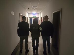 W korytarzu jednostki policji widziani z tyłu dwaj policjanci przytrzymujący mężczyznę