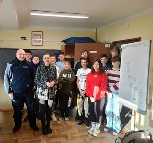 Grupa młodzieży wewnątrz sali szkolnej pozująca do zdjęcia z policjantem oraz wychowawczynią
