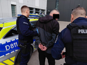 Mężczyzna ze skutymi z tyłu rękami przytrzymywany przez dwóch policjantów obok radiowozu na tle budynku jednostki