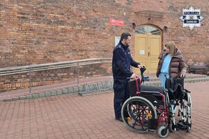 Na tle budynku z czerwonej cegły policjant trzymający wózek inwalidzki i kobieta w beżowej kurtce