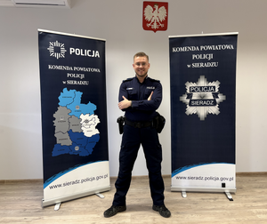 Uśmiechnięty policjant pomiędzy banerami Komendy Powiatowej Policji w Sieradzu