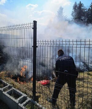 Za panelowym ogrodzeniem policjant z szuflą w kłębach dymu