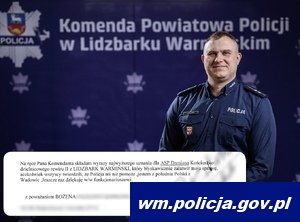 Policjant na tle granatowego baneru Komendy Powiatowej w Lidzbarku Warmińskim