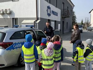Przed budynkiem policji policjant pokazujący grupce dzieci radiowóz policyjny