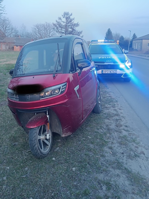 Na trawiastym poboczu drogi trójkołowiec, a za nim radiowóz