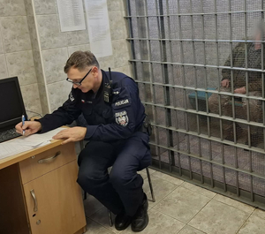 Policjant sporządzający przy biurku dokumentację, w drugim planie krata pomieszczenia przejściowego z umieszczonym wewnątrz mężczyzną