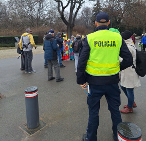 widziani od tyłu policjant w kamizelce i grupa starszych osób w alei parkowej