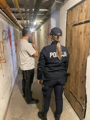 W korytarzu piwnicy budynku wielorodzinnego widziani z tyłu policjantka oraz mężczyzna