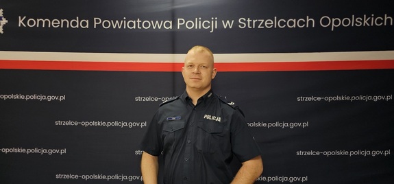 Uśmiechnięty policjant na tle banneru Komendy Powiatowej Policji w Strzelcach Opolskich