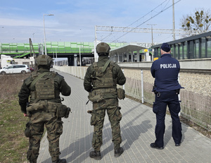 Widziani od tyłu policjant oraz dwaj żołnierze na chodniku obok przystanku kolejowego