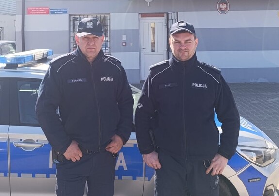 Dwaj policjanci na tle radiowozu i budynku komendy