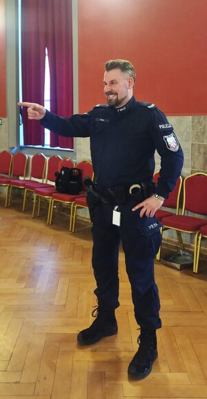 Stojący w sali uśmiechnięty policjant z wyciągniętą ręką i palcem wskazującym