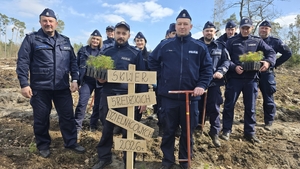 Kilkunastoosobowa grupa policjantek i policjantów z sadzonkami drzew i prowizorycznym drogowskazem "Skwer średzkich dzielnicowych 2026 r."