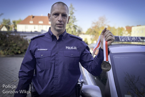 Policjant trzymający w ręku medal na biało-czerwonej szarfie  stojący obok radiowozu na tle rozmytego obrazu starówki