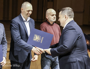 Mężczyzna w garniturze wręczający na scenie papierową teczkę drugiemu uśmiechniętemu mężczyźnie w garniturze, obok mężczyzna w swetrze