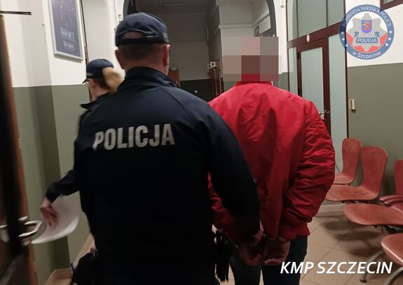 W korytarzu jednostki policji dwójka policjantów prowadząca mężczyznę ze skutymi kajdankami rękami