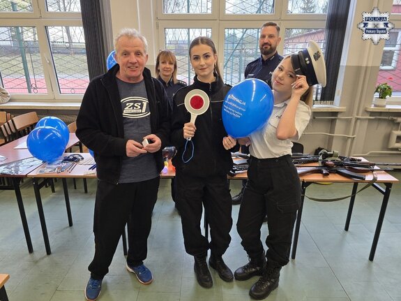 Pozujący do zdjęcia starszy mężczyzna, dwie nastolatki oraz w tle dwójka policjantów