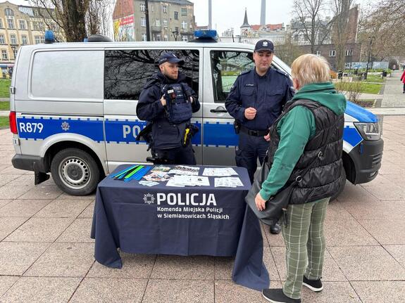 Na placu miasta dwóch policjantów ze stolikiem na tle radiowozu typu bus oraz seniorka