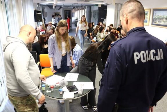 Przy szklanym okrągłym stoliku z różnego rodzaju fiolkami i kartami papieru policjant oraz mężczyzna ubrany w jasną bluzie, w tle grupa młodzieży