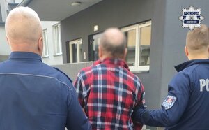 Widziani z tyłu dwaj policjanci prowadzący mężczyznę do budynku jednostki Policji
